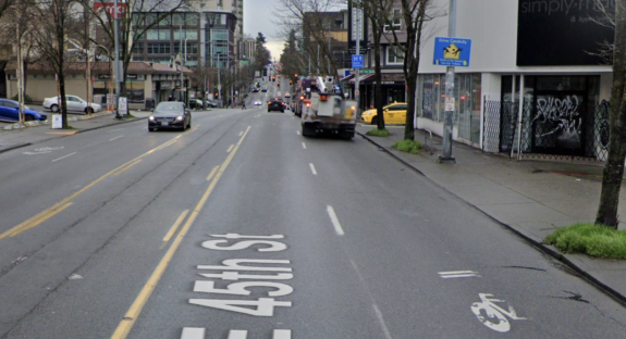 Photo from Google Street View of a mostly-erased sharrow in a mixed traffic lane on a five-lane street. A nearby sign says Drive Carefully and shows the stick figures from a pedestrian crossing sign comicaly diving out of the way.